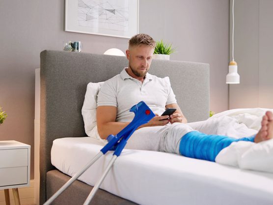 Mann mit Gipsbein sitzt daheim im Bett und sieht auf Smartphone