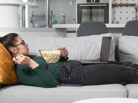 Frau liegt mit Popcorn und Laptop auf Sofa