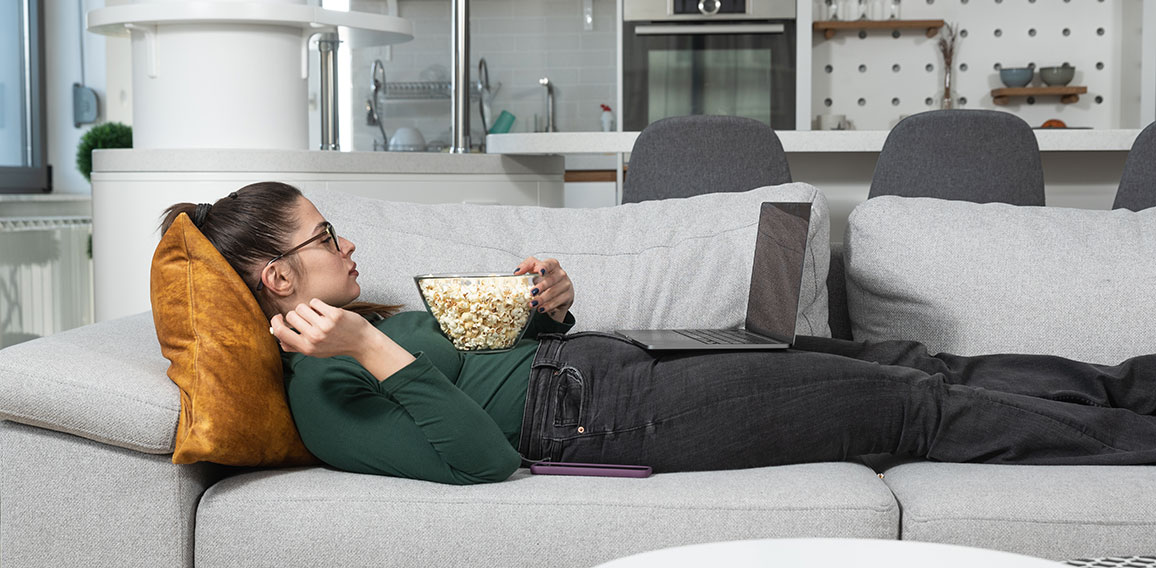 Frau liegt mit Popcorn und Laptop auf Sofa