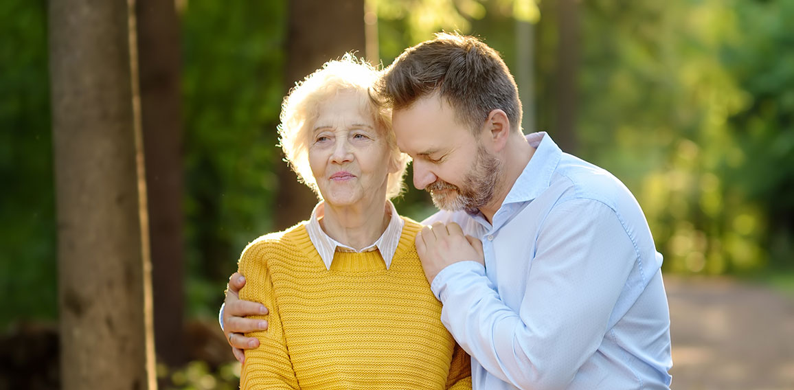 Loving adult son tenderly embracing his joyful elderly mother during walking at summer park. Mother's day holiday.