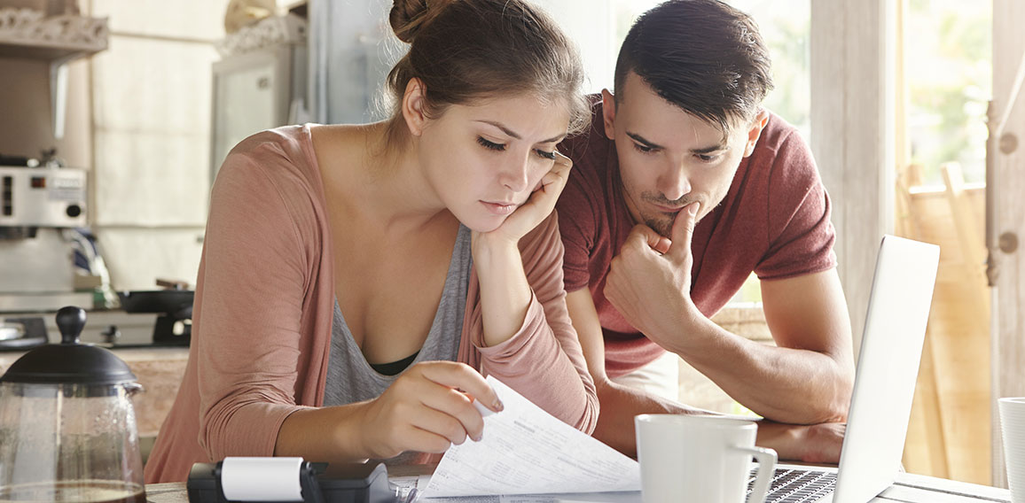 Worried young Caucasian married couple reading important notification from bank while managing domestic finances and calculating their expenses at kitchen table, using laptop computer and calculator