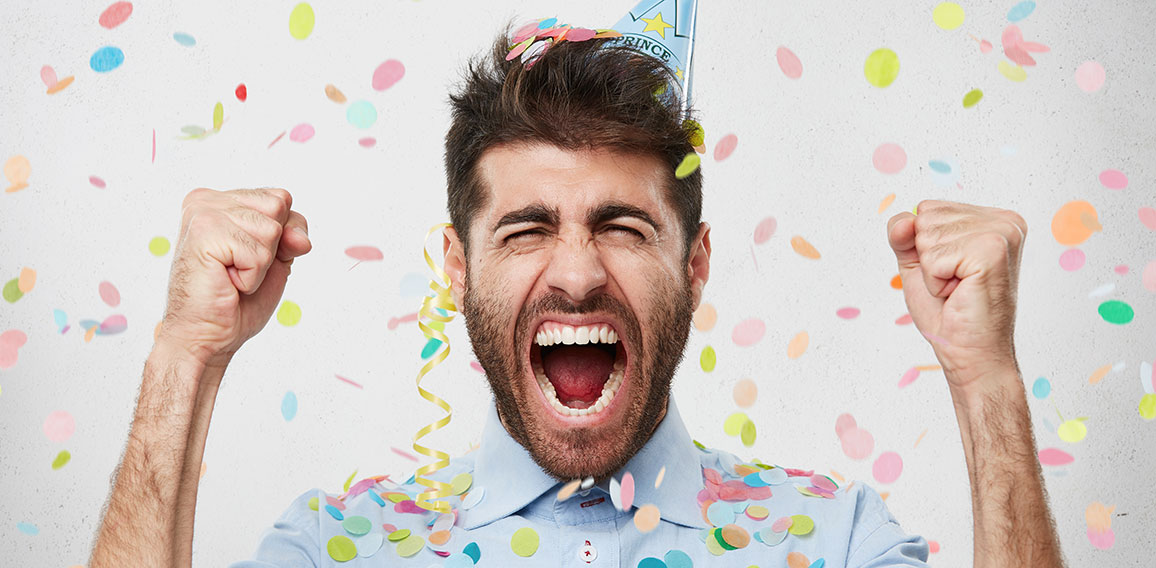 Joyful bearded man in formal clothes and holiday cap, clenching his fists with happiness, celebrating his birthday, having good mood. Satisfied male posing against white background with color pieces