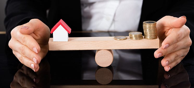 Model House And Money Coins Balancing On A Seesaw