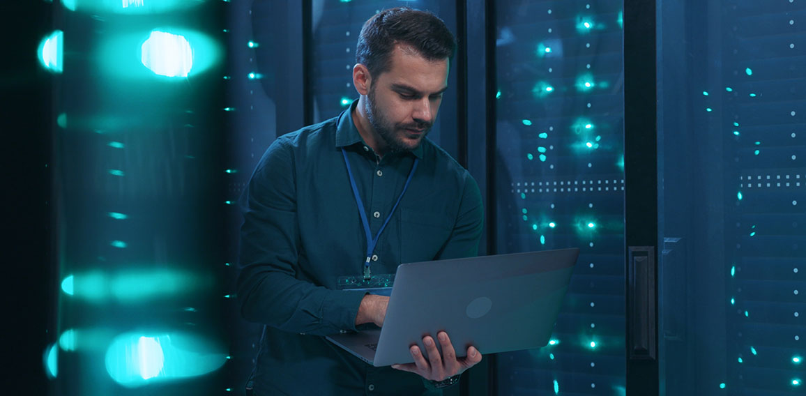 Caucasian IT professional admin using laptop computer doing data transfer operation with rack server cabinets in digital room of data center. Cyber security.