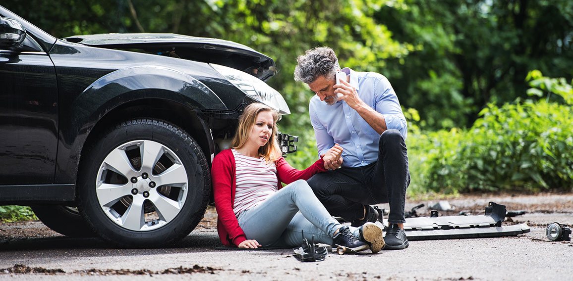 Young woman by the car after an accident and a man making a phone call.