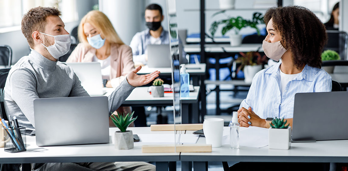 Communication and meeting in office after returning from quarantine. Young guy and african american woman in protective mask talking through glass board at workplace