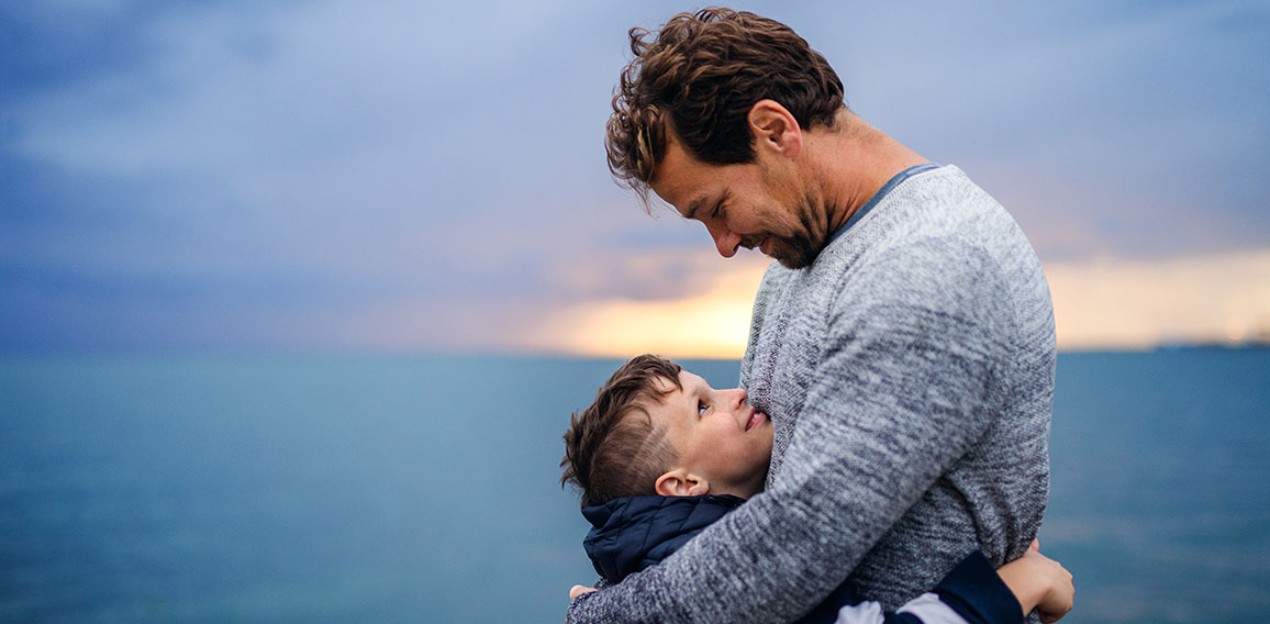 Father with small son on a walk outdoors standing on beach at dusk.