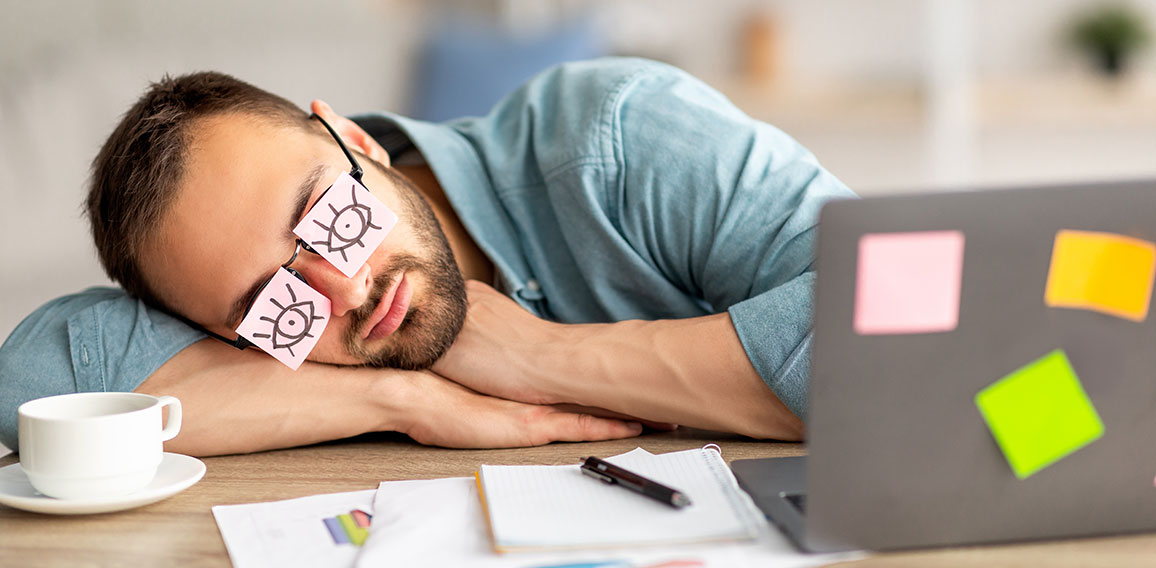 Lazy unproductive young guy wearing funny sticky notes with open eyes on his glasses, sleeping at workplace