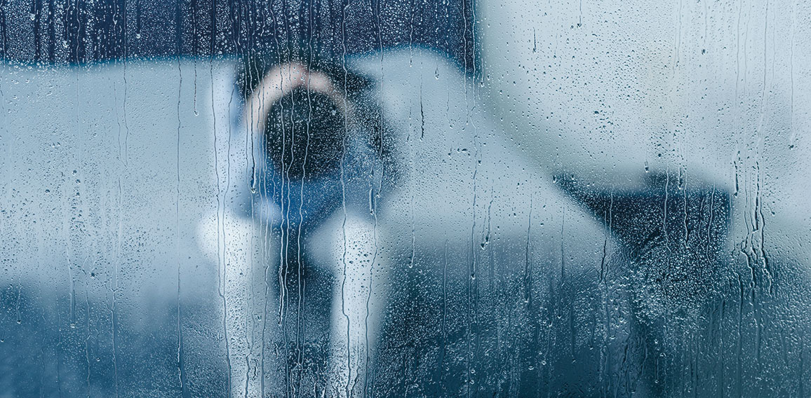 depressed woman sitting on bed and holding head in hands through window with raindrops