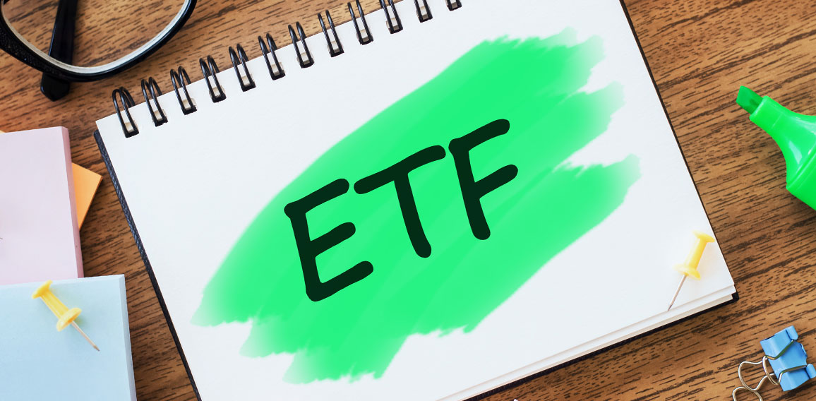 On the table are glasses, coloured markers, crumpled sheets, paper clips and a white notepad with text ETF Exchange Traded Funds highlighted in green. Business concept