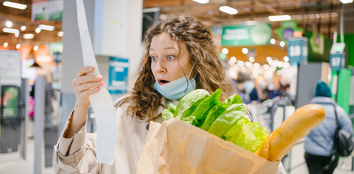 Young woman in a medical mask looks shocked at a paper check in a grocery supermarket holding a paper bag with groceries