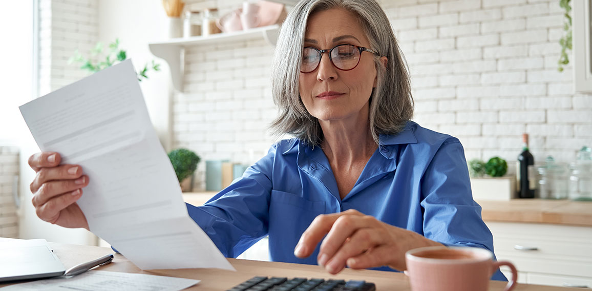 Mature woman holding paper bill using calculator managing finances at home.