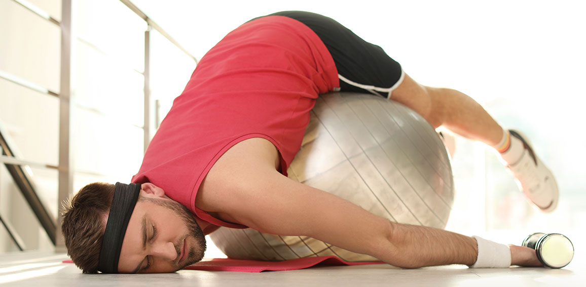 Lazy young man with exercise ball on yoga mat indoors