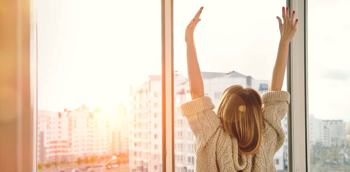 Woman near window raising hands facing the sunrise at morning