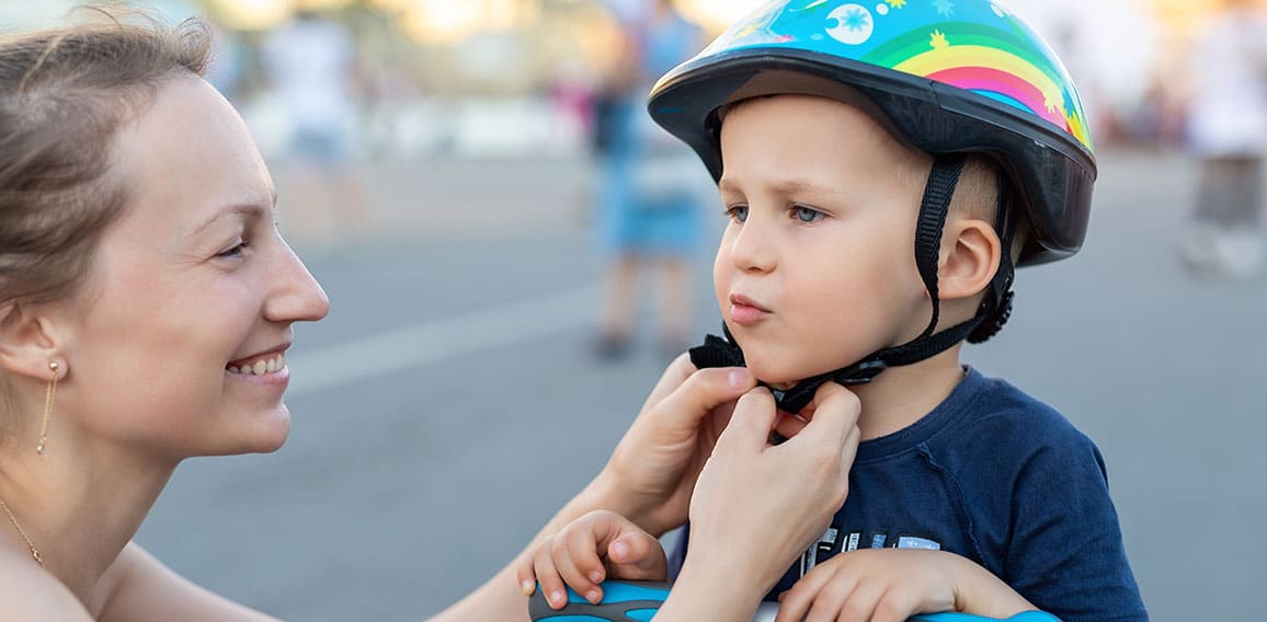 Close-up mom parent hand put on and fasten safety helmet on cute blond caucasian toddler boy for riding bike or scooter city street park outdoors on summer day. Child sport activity protection care