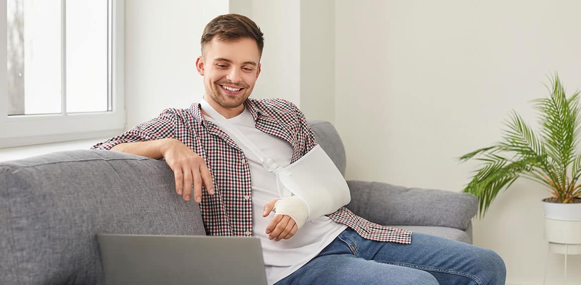 Happy young man with broken arm sitting on sofa at home and watching movie on laptop