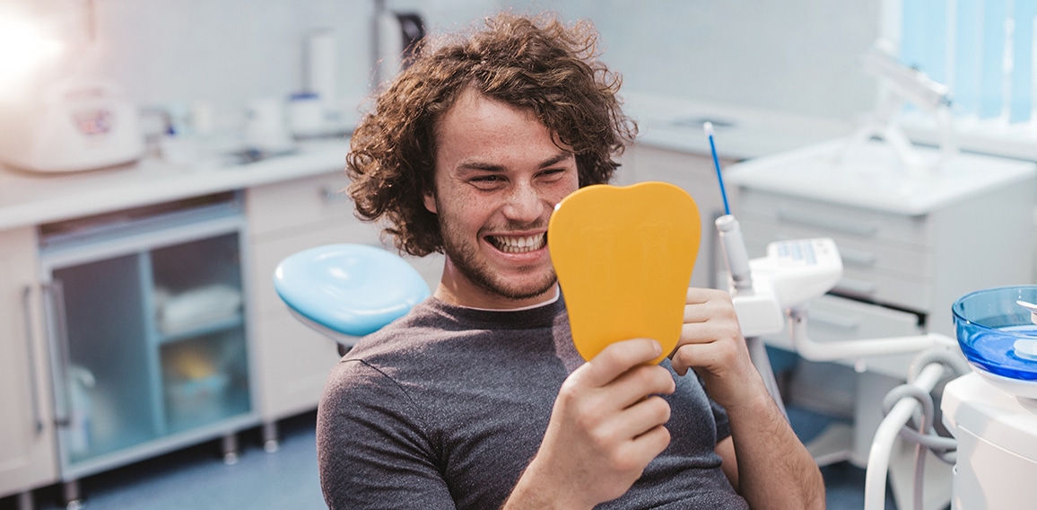 Charismatic smiling young man on the dentist chair , after a oral hygiene procedure take a mirror and looking at his white healthy teeth and are very happy for results , modern dental clinic