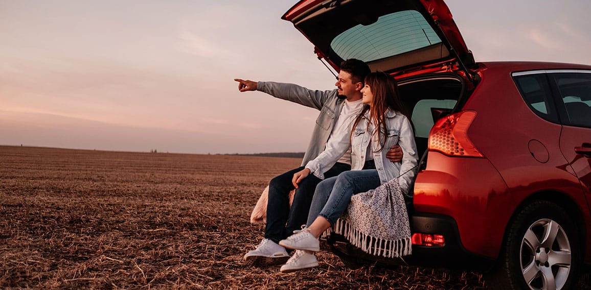 Young Happy Couple Dressed Alike in White Shirt and Jeans Sitting at Their New Car Trunk, Beautiful Sunset on the Field, Vacation and Travel Concept