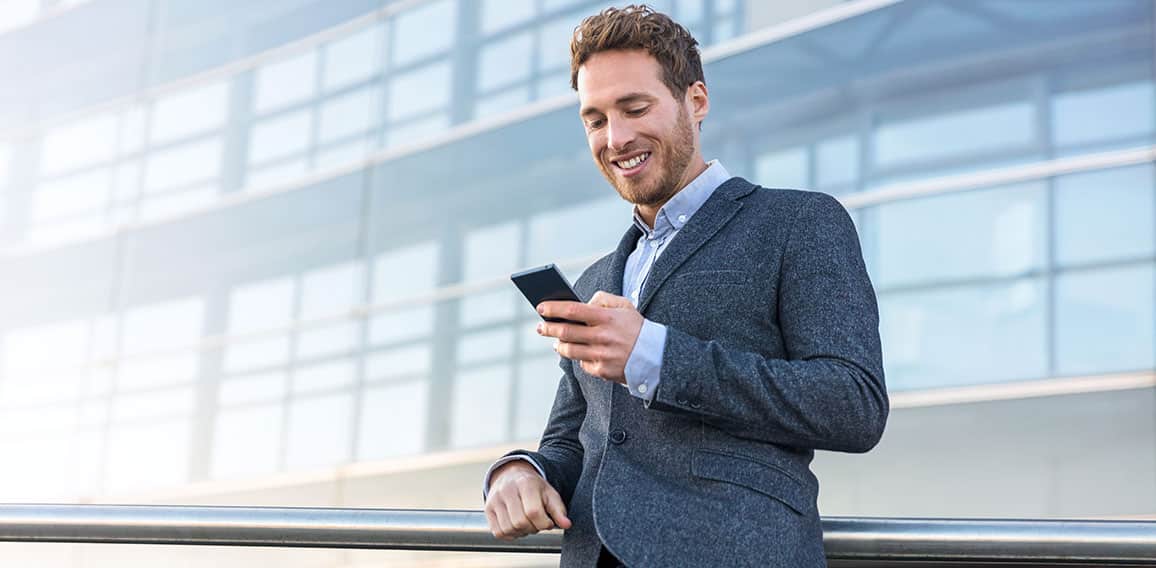 Businessman using mobile phone app texting outside of office in urban city with skyscrapers buildings in the background. Young caucasian man holding smartphone for business work.