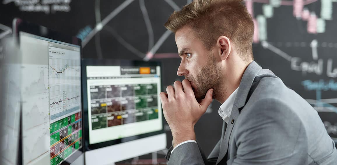 Keep growing with smart ideas. Portrait of successful young trader looking focused while sitting in front of multiple monitors in the office. Blackboard full of chart and data analyses in background.