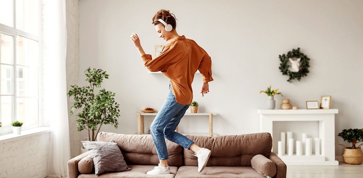 Cheerful woman listening to music and dancing on soft couch at home in day off