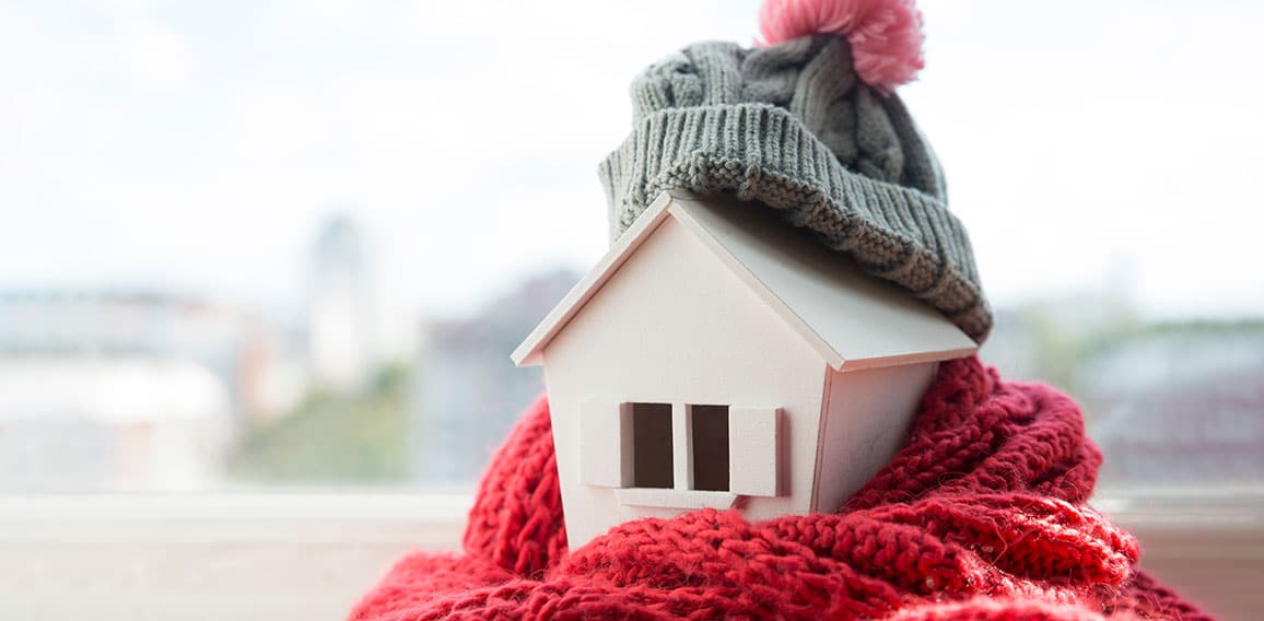 house in winter - heating system concept and cold snowy weather with model of a house wearing a knitted cap