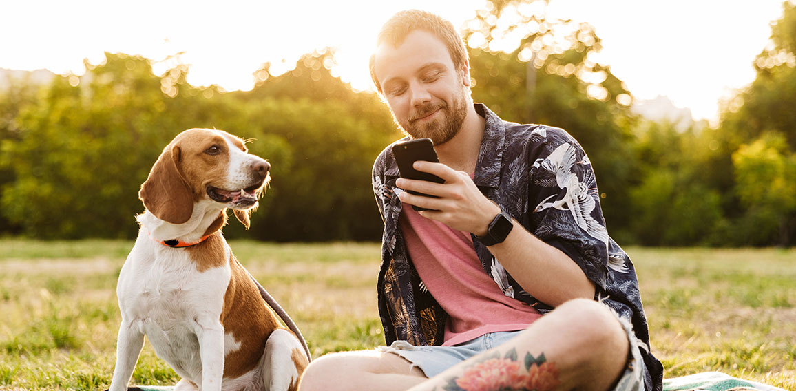 Image of young man using cellphone and sitting with beagle dog in park