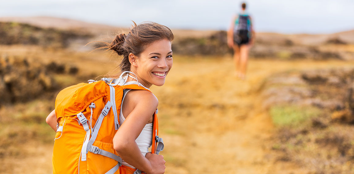 Happy young Asian hiker girl hiking with friend in mountain nature trail wearing orange backpack smiling looking back enjoying travel holiday in summer. Adventure wanderlust lifestyle. Healthy woman.