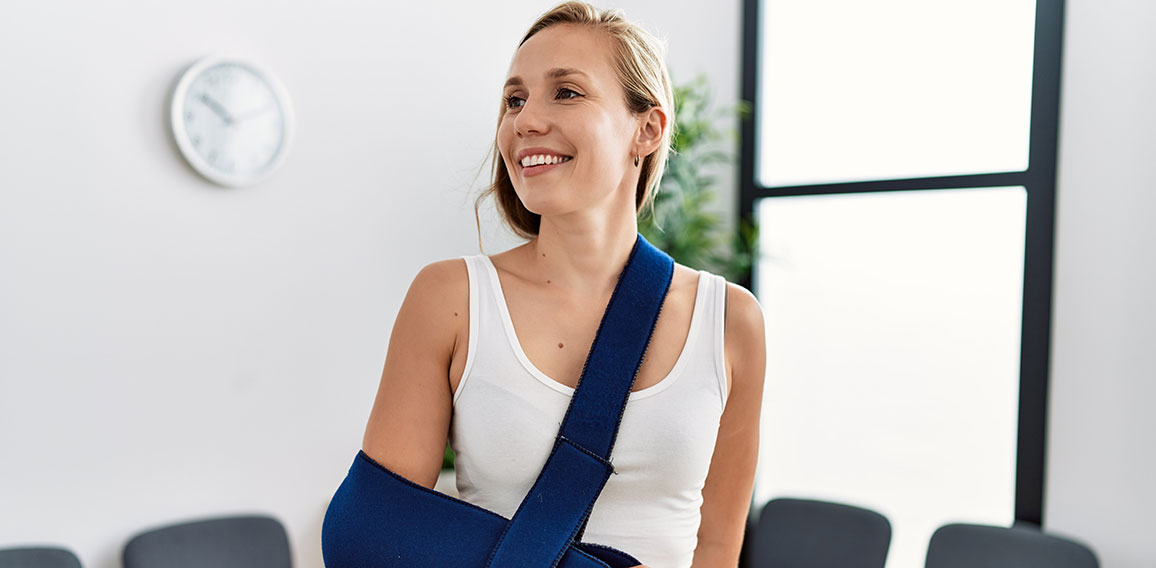 Young caucasian woman wearing sling on arm for accident standing at clinic waiting room