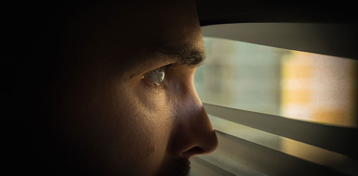 A man in a dark room looks out the window through the blinds. Covert surveillance. Close-up.