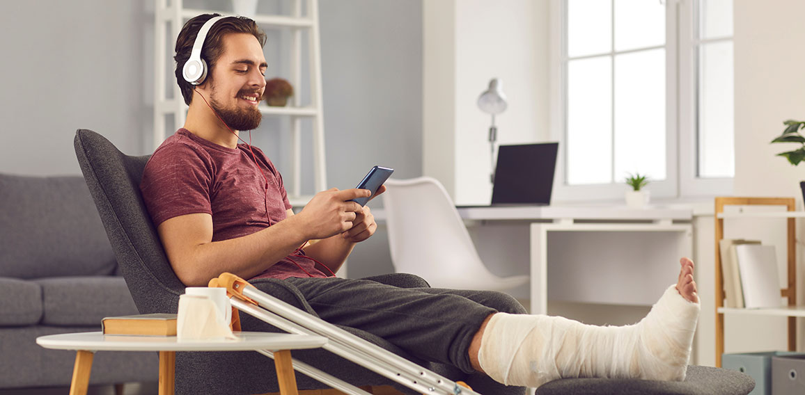 Young man with broken leg sitting in chair and listening to music on mobile phone