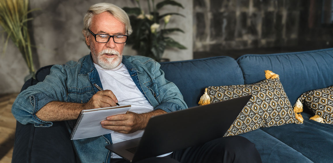 Focused elderly man writing notes in notebook watching webinar or online training using laptop computer modern senior male with gray hair and beard learning online