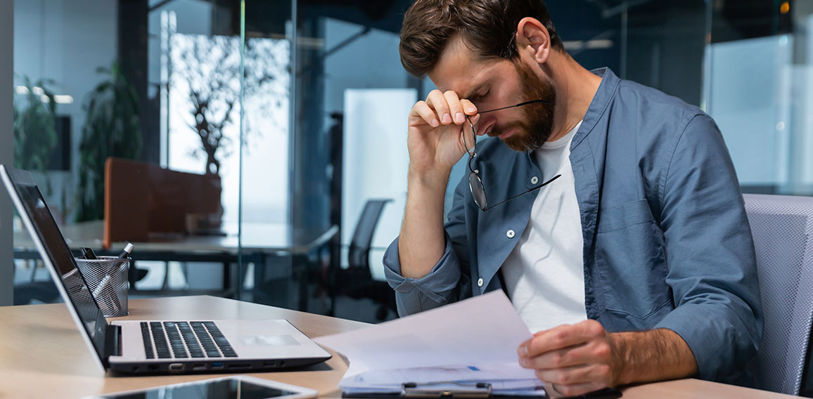 Upset businessman behind paper work inside modern office, mature man with beard reading financial reports and account documents unhappy with results and disappointed with achievements