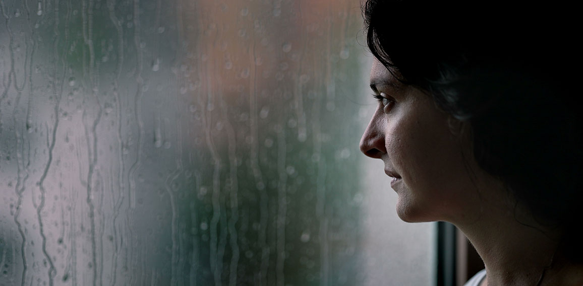 Woman standing by window during rainy day looking outside watchi