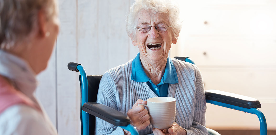 Senior woman, friend and coffee or tea while in a wheelchair for a disability or rehabilitation and happy or laughing about funny conversation or joke. Old people together for fun in nursing home