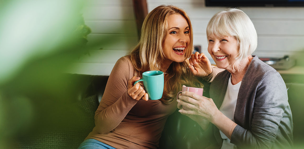 Happy senior mother with adult daughter sitting on couch and holding cups with coffee or tea at home. Togetherness concept