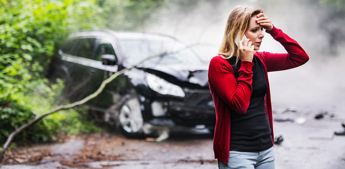 A young woman with smartphone by the damaged car after a car accident, making a phone call.