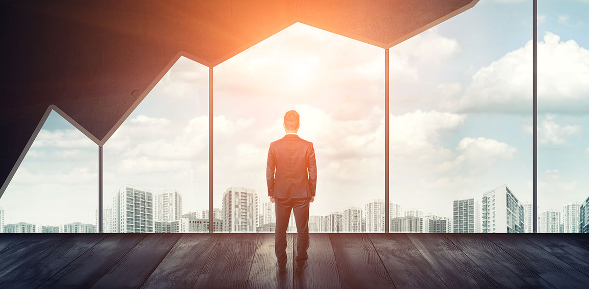 Businessman standing on the floor back to us looking through the big window with cityscape and sunset behind it.