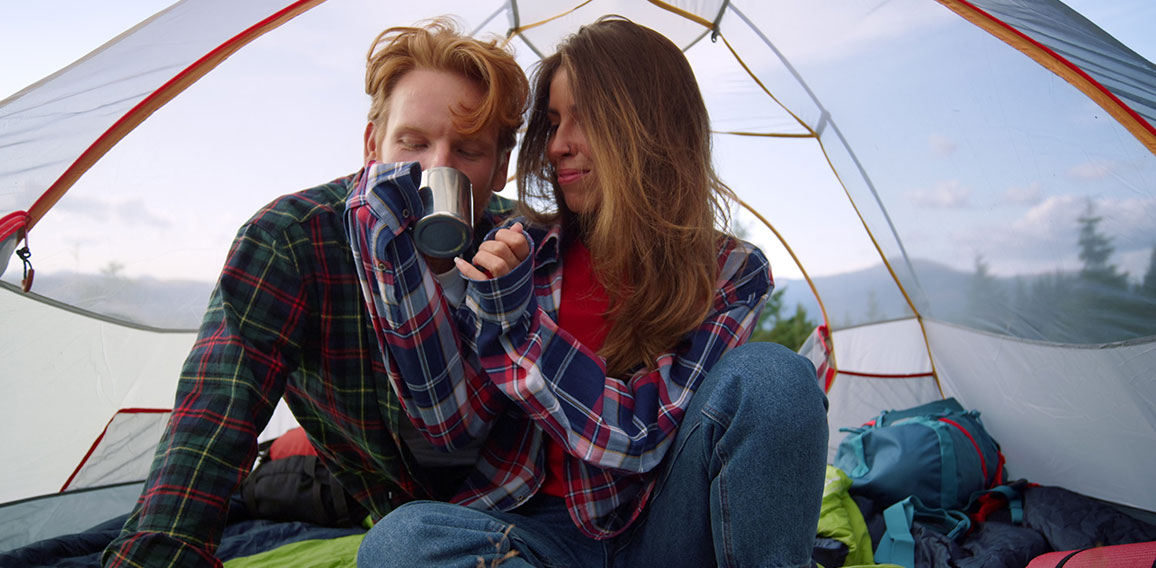 Young couple sitting in tent during hike. Redhead guy drinking tea from cup