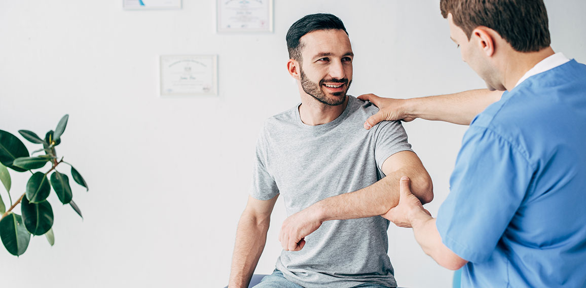 Smiling patient sitting on couch and doctor examining patient shoulder in massage cabinet at clinic