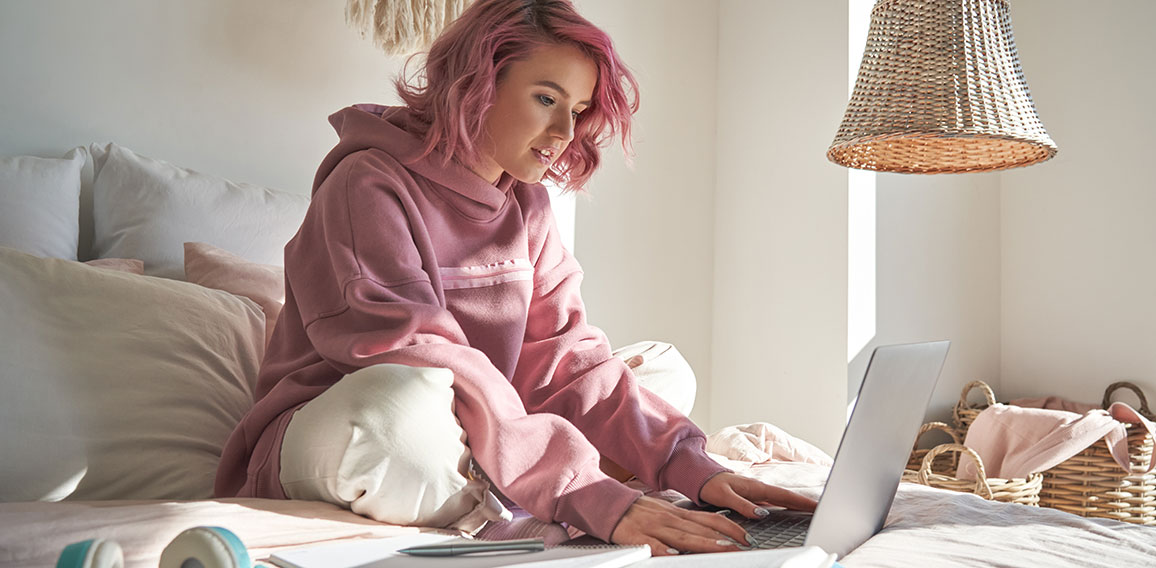 Hipster teen girl student with pink hair using laptop online learning in bed.