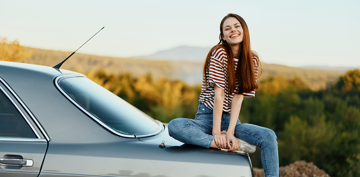 A woman car driver sits on the trunk of a car and smiles admiring a beautiful view of autumn nature and mountains