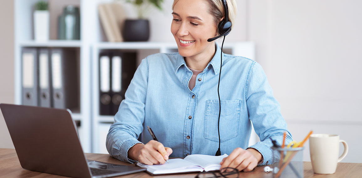 Happy young woman in headset participating in online lesson or web conference, taking notes, studying remotely