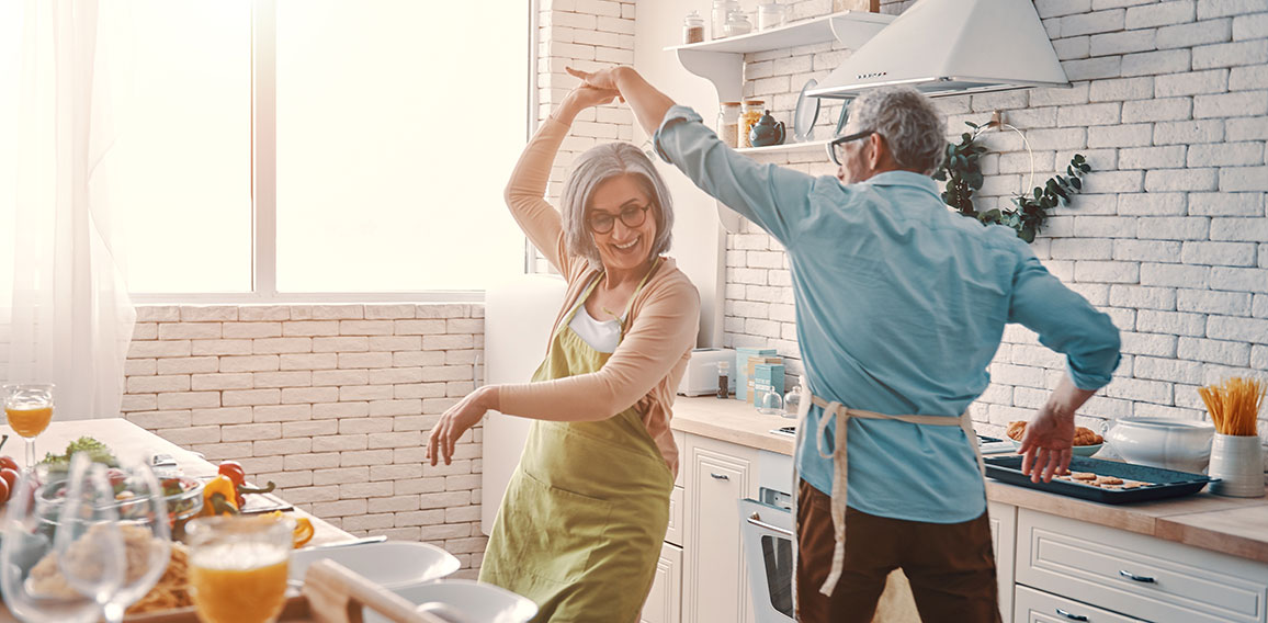 Beautiful playful senior couple in aprons dancing and smiling while preparing healthy dinner at home