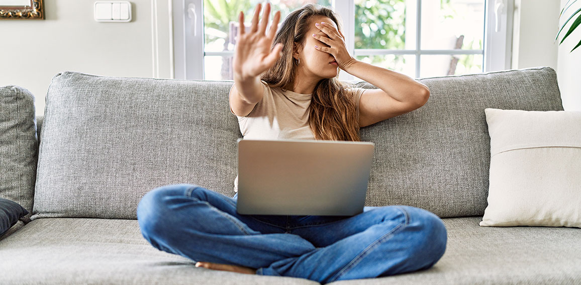 Beautiful young brunette woman sitting on the sofa using computer laptop at home covering eyes with hands and doing stop gesture with sad and fear expression. embarrassed and negative concept.