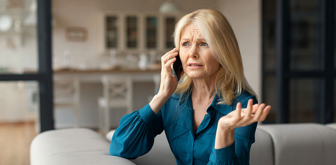 Stressed mature woman talking on cellphone, having unpleasant phone conversation while sitting on sofa at home