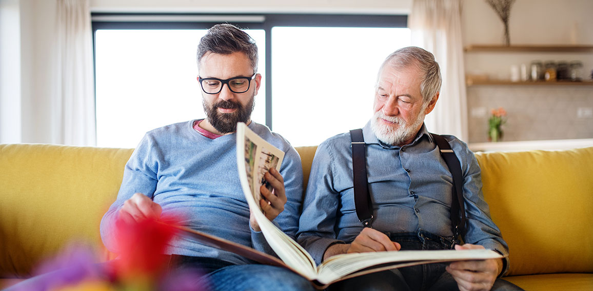 An adult hipster son and senior father sitting on sofa indoors at home.