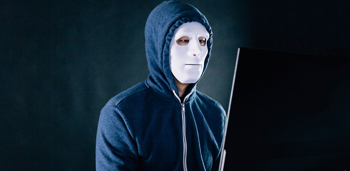 Anonymous and masked hacker under hoodie using computer isolated over dark background - illegal online internet criminal concept