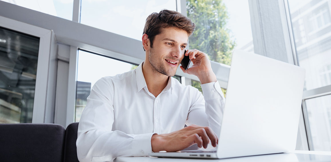 Happy young businessman talking on cell phone and using laptop