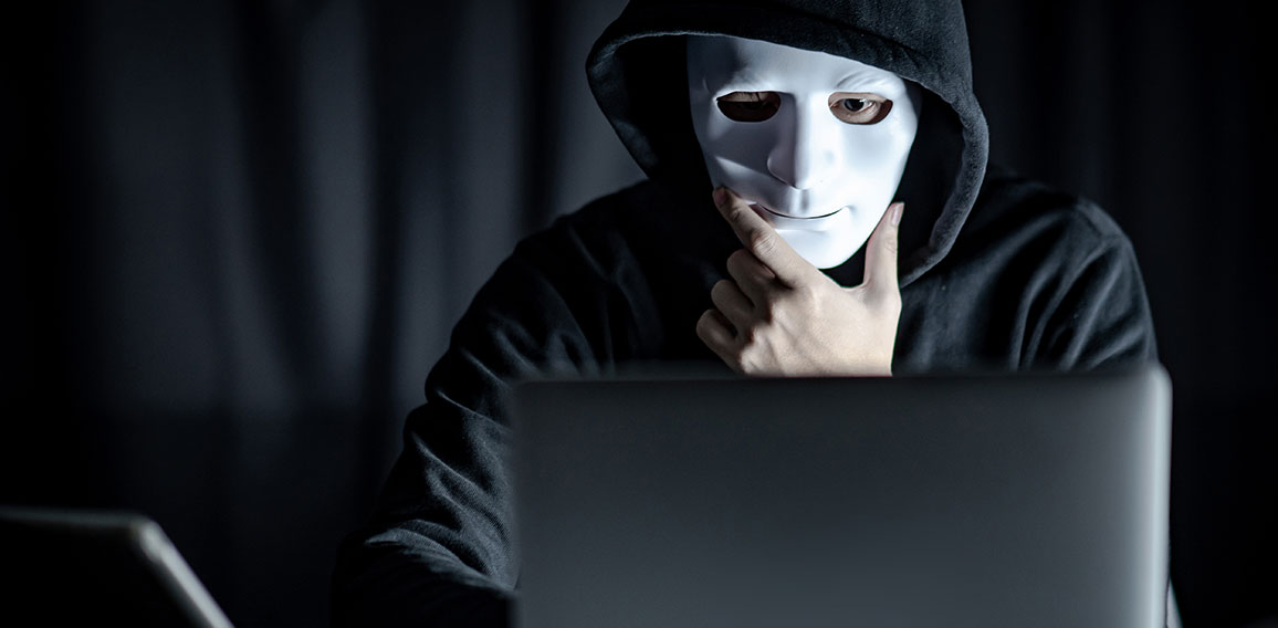 Mystery male hoodie hacker holding white mask looking at laptop